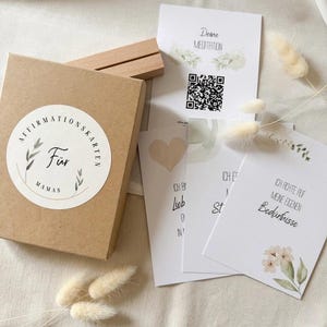 May include: A kraft paper box with a round sticker that reads "Affirmationskarten Für Mamas". Several white cards with floral accents and German text are scattered around the box. Dried flowers and a wooden card holder are also present.
