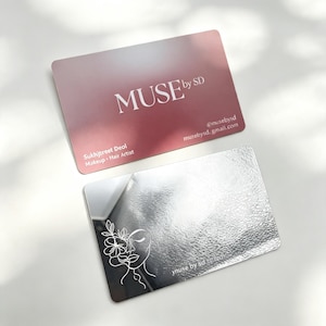 May include: Two rectangular business cards. One card is a matte dusty rose color with white text reading MUSE by SD, Sukhjpreet Deol Makeup Hair Artist, and contact details. The second card is a reflective silver metallic with a white line art face.