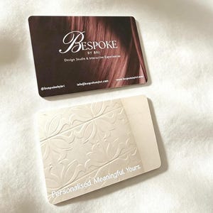 May include: Two rectangular business cards. The top card is dark brown with the white text Bespoke by Bri, Design Studio & Interactive Experiences, an email address, and a website. The bottom card is cream with an embossed floral tile pattern.