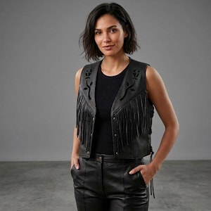May include: Black leather vest featuring fringe detailing and floral cutouts. The vest has a V-neck, button closure, and side lacing. It is worn over a black top and paired with black leather trousers. The style evokes a biker or western aesthetic.