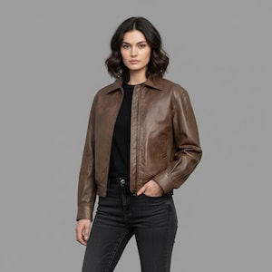 Puede incluir: Chaqueta de cuero marrón con cuello y cierre de cremallera. La chaqueta tiene mangas largas y un estilo bomber clásico. La modelo lleva una camiseta negra y vaqueros grises oscuros.