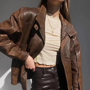 May include: A brown leather jacket with a wide collar and a button closure. The jacket is worn over a cream-colored top and dark brown pants. A gold necklace is visible. The jacket appears to be vintage or retro style.
