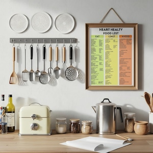 May include: A kitchen scene with a variety of cooking utensils hanging above a countertop. A framed "Heart Healthy Food List" hangs on the wall. A cream-colored appliance, jars, and a stainless steel kettle are on the counter.