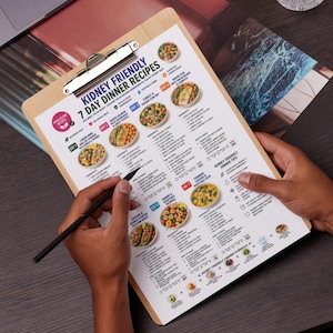 May include: A wooden clipboard holding a printed document titled Kidney Friendly 7 Day Dinner Recipes. The page features a structured meal plan with daily food illustrations, ingredient lists, and cooking instructions for seven distinct dinner meals.