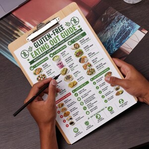 K&ouml;nnte beinhalten: Ein h&ouml;lzerner Klemmbrett mit einem gedruckten "Gluten-Free Eating Out Guide" wird gehalten. Der Leitfaden listet Restaurant-, Caf&eacute;- und Fast-Food-Bestellideen auf, einschlie&szlig;lich Lebensmittelaustausch und sicherer Optionen. Ein schwarzer Stift zeigt auf den Leitfaden.