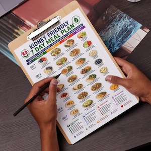 May include: A wooden clipboard holding a printed Kidney Friendly 7 Day Meal Plan chart. The document features a grid of daily breakfast, lunch, and dinner meal suggestions with small food illustrations, nutritional guidelines, and tips for success.