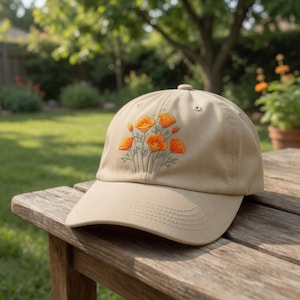 May include: A tan baseball cap with an embroidered design of orange poppies and green stems. The cap is sitting on a wooden surface, with a blurred green background of trees and grass.