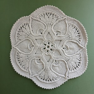 Könnte beinhalten: Ein rundes, hellbeiges Häkeldeckchen mit einem komplexen, symmetrischen Blumenmuster aus Reliefmaschen. Das Design strahlt von einem zentralen Ring aus und zeigt filigrane blattartige Motive sowie einen gewellten, strukturierten Rand.