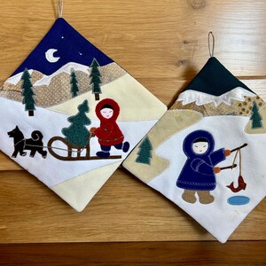 May include: Two white quilted pot holders with winter scenes. One features a person in a red coat pulling a sled with a Christmas tree. The other features a person in a blue coat fishing.