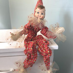 May include: A vintage red and white fabric doll dressed as a jester with a pointed hat, white ruffles, and a yellow flower pattern. The doll is sitting on a white dresser.