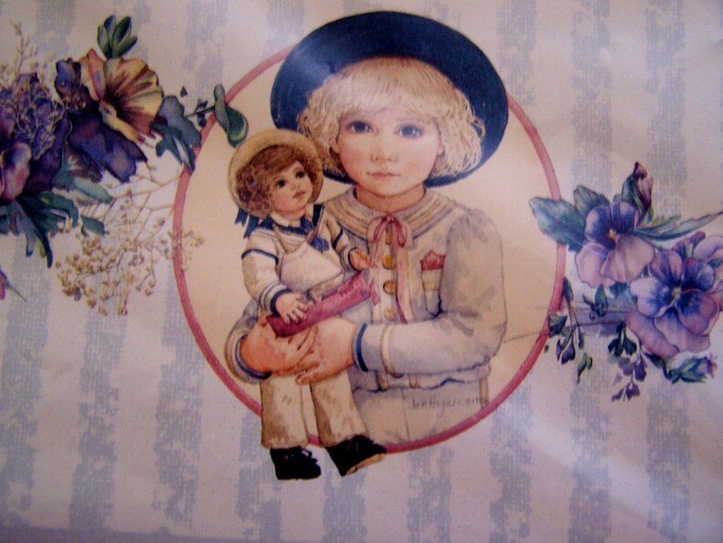 Pode incluir: Uma ilustra&ccedil;&atilde;o vintage de uma menina vestindo um traje de marinheiro azul e um chap&eacute;u preto segurando uma boneca com um vestido branco e um colar de marinheiro azul. A ilustra&ccedil;&atilde;o &eacute; cercada por um c&iacute;rculo rosa e detalhes florais.
