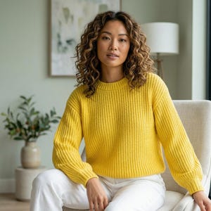 Puede incluir: Una mujer con un suéter de punto amarillo brillante con textura acanalada y mangas largas. El suéter se combina con pantalones blancos. La mujer está sentada en una silla de color claro en una habitación bien iluminada.