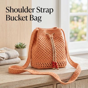 Puede incluir: Un bolso de cubo de ganchillo naranja con una correa para el hombro. El bolso tiene un cierre de cordón y un cordón decorativo con cuentas rojas. El texto "Shoulder Strap Bucket Bag" está en la parte superior de la imagen.