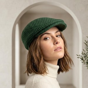 May include: A dark green newsboy cap with a textured pattern. The cap has a small brim and is worn by a person wearing a cream-colored turtleneck sweater.