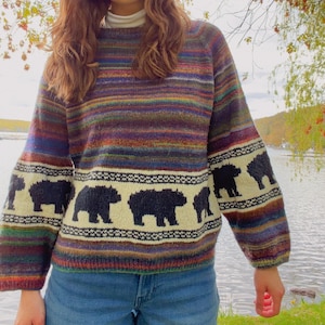 May include: A long-sleeved knit sweater featuring a horizontal multi-colored striped pattern in shades of purple, blue, green, and orange. A cream-colored band across the chest and sleeves displays a repeating black bear silhouette motif.