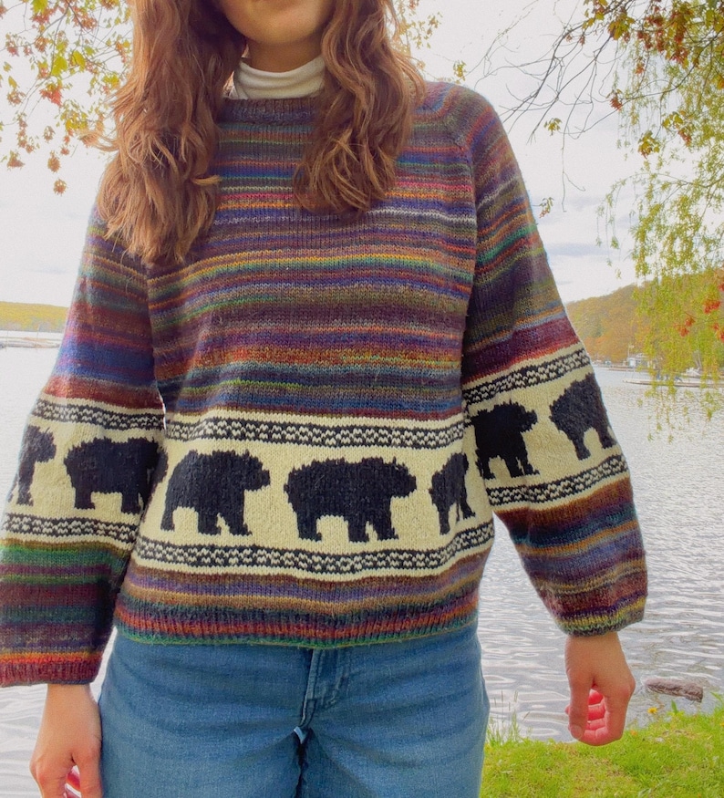 May include: A long-sleeved knit sweater featuring a horizontal multi-colored striped pattern in shades of purple, blue, green, and orange. A cream-colored band across the chest and sleeves displays a repeating black bear silhouette motif.
