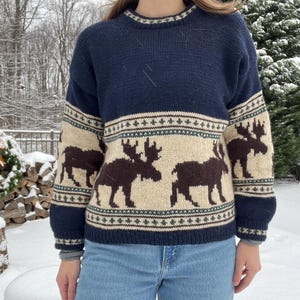 May include: Navy blue knit sweater with a cream-colored band featuring brown moose silhouettes. The sweater has a crew neck and ribbed cuffs. The design includes green and white geometric patterns. A cozy winter garment.