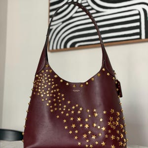 May include: A burgundy leather shoulder bag with gold star-shaped studs. The bag has a single shoulder strap and a curved shape. The word "COACH" is embossed on the front. The bag is on a marble surface.