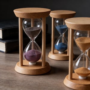 May include: Three wooden hourglasses with clear glass bulbs filled with colored sand: purple, blue, and orange. The hourglasses have a light wood frame and base. The background includes a dark surface and a stack of books.