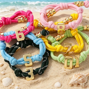 Op de afbeelding: Een verzameling kleurrijke touwarmbanden met goudkleurige nummerbedels. De armbanden zijn in tinten roze, blauw, groen en geel, met gouden accenten. De armbanden liggen op zand in de buurt van de zee.