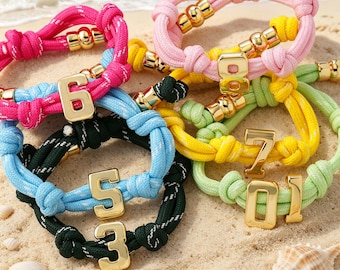 May include: A collection of colorful rope bracelets with gold-colored number charms. The bracelets are in shades of pink, blue, green, and yellow, with gold accents. The bracelets are displayed on a sandy surface near the ocean.