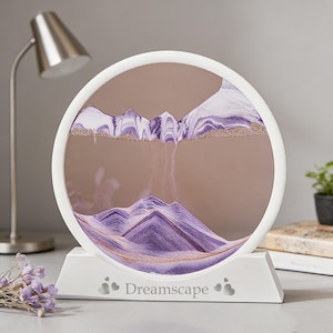 Puede incluir: Una pieza de arte de arena enmarcada en blanco con un diseño circular. El arte de arena presenta un paisaje montañoso en tonos morado, blanco y dorado. La base del marco es blanca y tiene la palabra "Dreamscape" impresa.