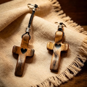 May include: Two wooden cross keychains with a heart cutout and engraved names. The crosses are attached to a black cord and silver-colored metal ring. The keychains are made of a light brown wood with a visible wood grain.