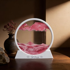 Peut inclure: Une pièce d'art de sable circulaire blanche avec du sable rose et doré, ressemblant à un paysage de montagne. La base indique "Soft Eternity". L'art du sable est présenté dans un cadre et une base blancs, créant un objet décoratif unique.