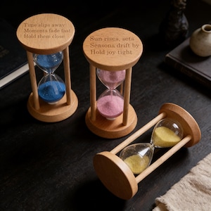 May include: Three wooden hourglasses with different colored sand: blue, pink, and yellow. The hourglasses have text engraved on the top, including "Time slips away, Moments fade fast, Hold them close" and "Sun rises, sets, Seasons drift by, Hold joy tight."