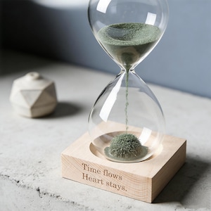 Puede incluir: Reloj de arena de cristal transparente con arena verde, sobre una base de madera clara. La base tiene el texto grabado "Time flows Heart stays." Un pequeño objeto decorativo blanco y geométrico está en el fondo. El reloj es decorativo.