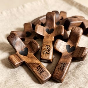 May include: Several wooden crosses with a heart-shaped cutout and personalized names. The crosses are made of dark wood with a visible grain. The names "Elsa", "Sarah", and "Millie" are engraved on the crosses.