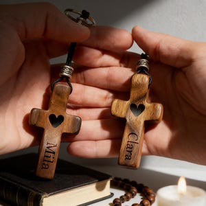 May include: Two wooden cross keychains with heart cutouts and personalized names. The crosses are brown with a dark heart-shaped cutout. One keychain says "Mila" and the other says "Clara". The keychains are held in hands.
