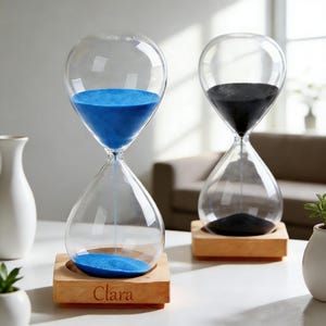 May include: Two clear glass hourglasses with wooden bases. One contains blue sand, the other black. The base of the blue hourglass is engraved with the name "Clara". The hourglasses are on a white surface.