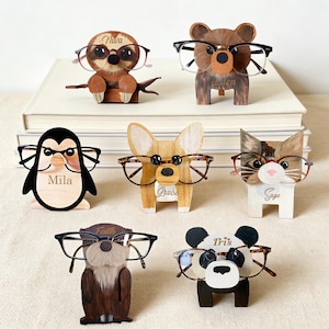 Puede incluir: Una colección de soportes de gafas de madera con forma de animales. Los soportes incluyen un perezoso, un oso, un pingüino, un perro, un gato, una nutria y un panda, cada uno con gafas. Los nombres están grabados en algunos de los soportes.