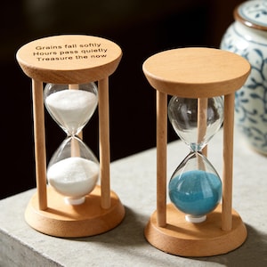 May include: Two wooden hourglasses with clear glass bulbs. One contains white sand, the other blue sand. The top of one hourglass reads "Grains fall softly, Hours pass quietly, Treasure the now."