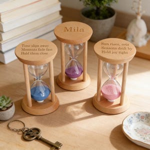 May include: Three wooden hourglasses with clear glass bulbs and colored sand: blue, purple, and pink. The top of each hourglass has an engraved message. One says "Mila". The other two read "Time slips away..." and "Sun rises, sets..."