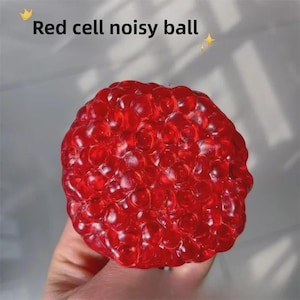 Pode incluir: Uma bola vermelha vibrante e texturizada com aparência borbulhante. O texto "Red cell noisy ball" é exibido no topo. A bola é segurada em uma mão contra um fundo cinza claro.