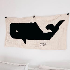 May include: A beige wall hanging with a large black whale design. The whale is geometric, with the text "A WHALE PASSES" below it. The hanging is secured with clips and hangs above a bed with striped pillows.