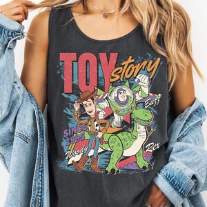 May include: Dark gray tank top featuring a graphic of characters from the movie "Toy Story." The design includes Woody, Buzz Lightyear, and Rex, with the words "Toy Story" and "Since 1995." The tank top has a vintage, distressed look.