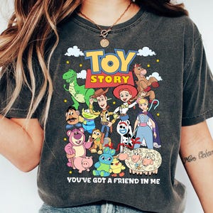 May include: Dark gray t-shirt featuring a colorful graphic of characters from the animated film "Toy Story." The design includes the film's logo and the phrase "You've Got a Friend in Me."