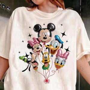 May include: Cream-colored t-shirt featuring a graphic of Mickey Mouse, Minnie Mouse, Donald Duck, Daisy Duck, and Goofy as balloon characters. The balloons are connected by red strings, with black star accents. The shirt is a casual style.