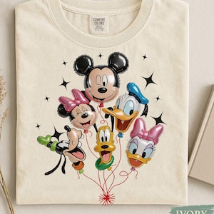 May include: Ivory-colored t-shirt featuring a graphic of Mickey Mouse, Minnie Mouse, Donald Duck, Daisy Duck, and Goofy as balloon characters. The balloons are connected by red strings, with black star accents. The shirt has a crew neck and short sleeves.