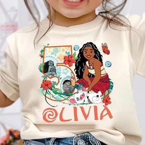 May include: A cream-colored t-shirt featuring a colorful graphic of a cartoon character with long black hair, a red skirt, and a necklace. The graphic includes the number 5, flowers, and the name "Olivia" in red. The shirt is worn by a young child.