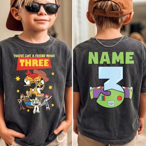 May include: Dark gray t-shirt featuring Toy Story characters and the text "YOUVE GOT A FRIEND WHOS THREE" on the front. The back of the shirt has the word "NAME" above a large number "3" with a Buzz Lightyear design.