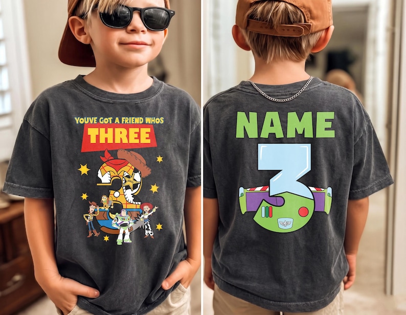 May include: Dark gray t-shirt featuring Toy Story characters and the text "YOUVE GOT A FRIEND WHOS THREE" on the front. The back of the shirt has the word "NAME" above a large number "3" with a Buzz Lightyear design.