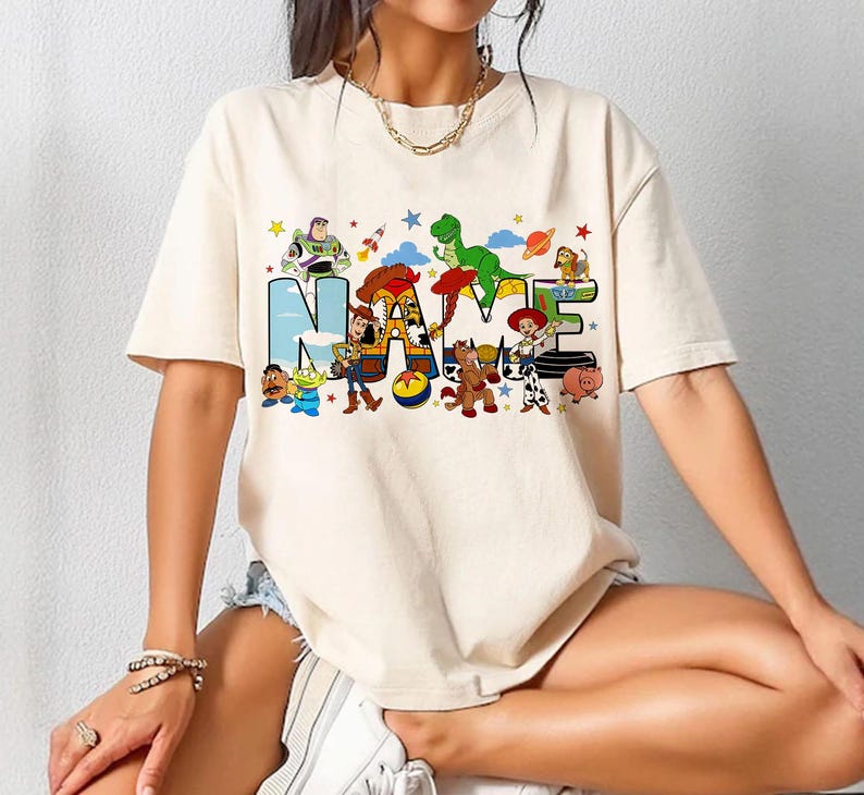 May include: Cream-colored t-shirt featuring the word "NAME" in large, colorful letters with cartoon characters. The design includes characters like Buzz Lightyear, Woody, and Rex, with stars and other playful elements.