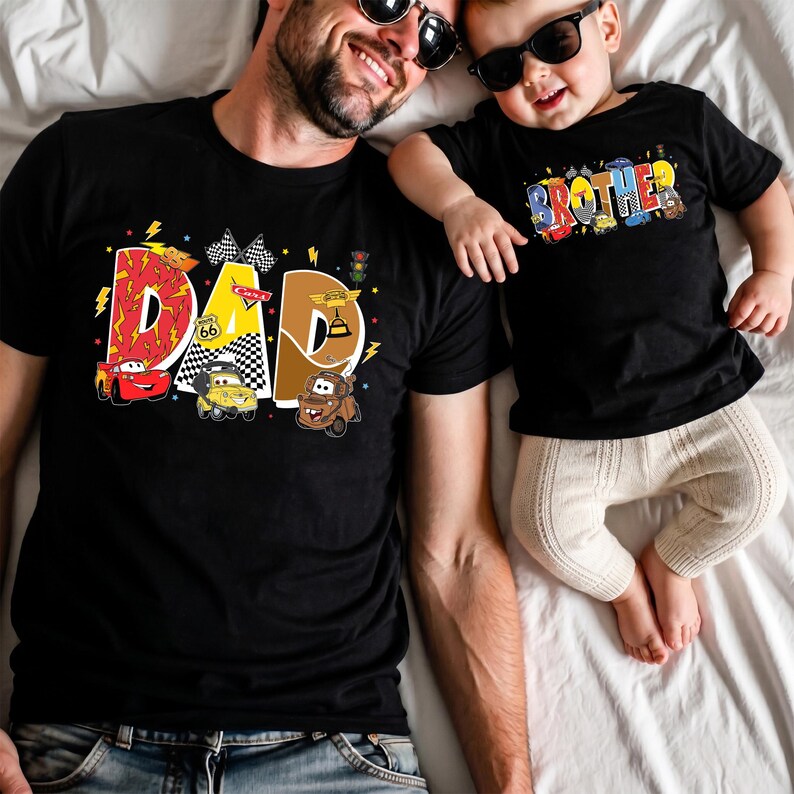 Może przedstawiać: Czarne koszulki z kolorową grafiką. Jedna koszulka m&oacute;wi "DAD" z ilustracjami samochod&oacute;w, druga m&oacute;wi "BROTHER" z podobnymi wzorami samochod&oacute;w. Obie koszulki mają motyw Cars i są noszone przez ojca i dziecko.