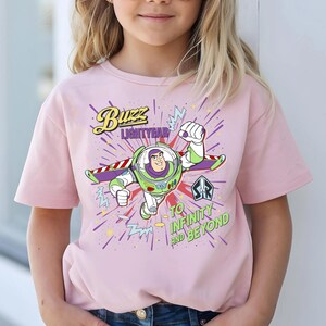 May include: A pale pink t-shirt with a graphic of Buzz Lightyear. The design includes the text "Buzz Lightyear" and "To Infinity and Beyond" with a cartoon image of the character in a flying pose.