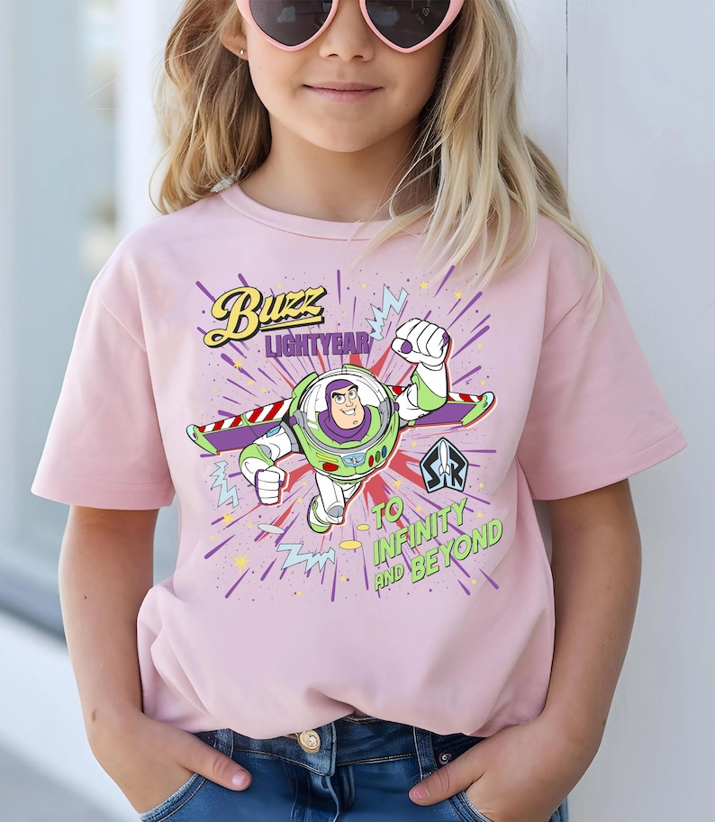 May include: A pale pink t-shirt with a graphic of Buzz Lightyear. The design includes the text "Buzz Lightyear" and "To Infinity and Beyond" with a cartoon image of the character in a flying pose.