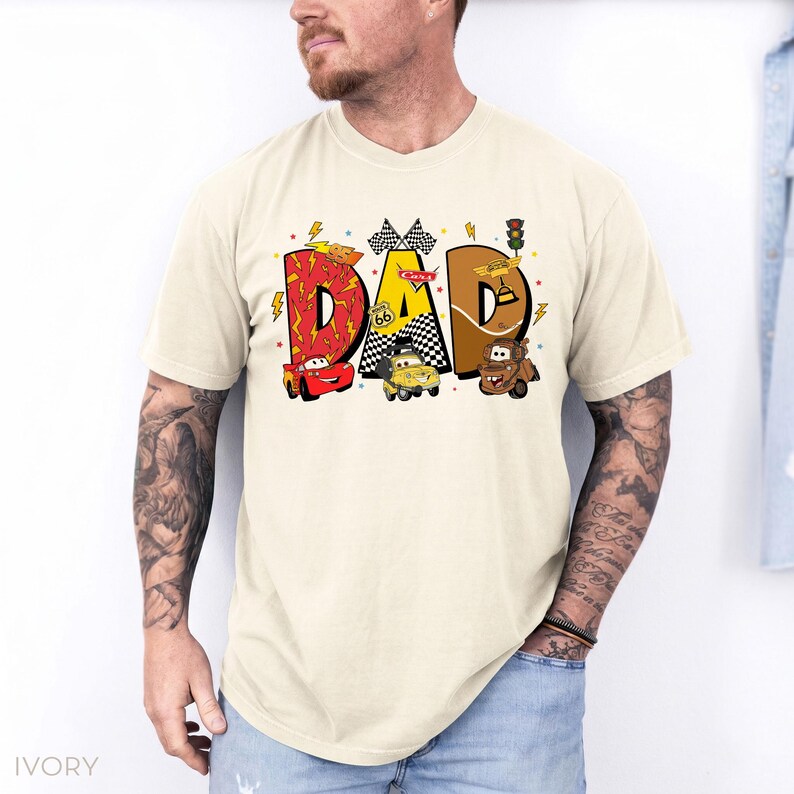 Może przedstawiać: Kremowa koszulka z napisem "DAD" w kolorowym, kresk&oacute;wkowym wzorze. Litery wypełnione są grafiką o tematyce wyścigowej, w tym samochodami, szachownicami i obrazami Route 66. Koszulka jest w stylu casual z kr&oacute;tkim rękawem.